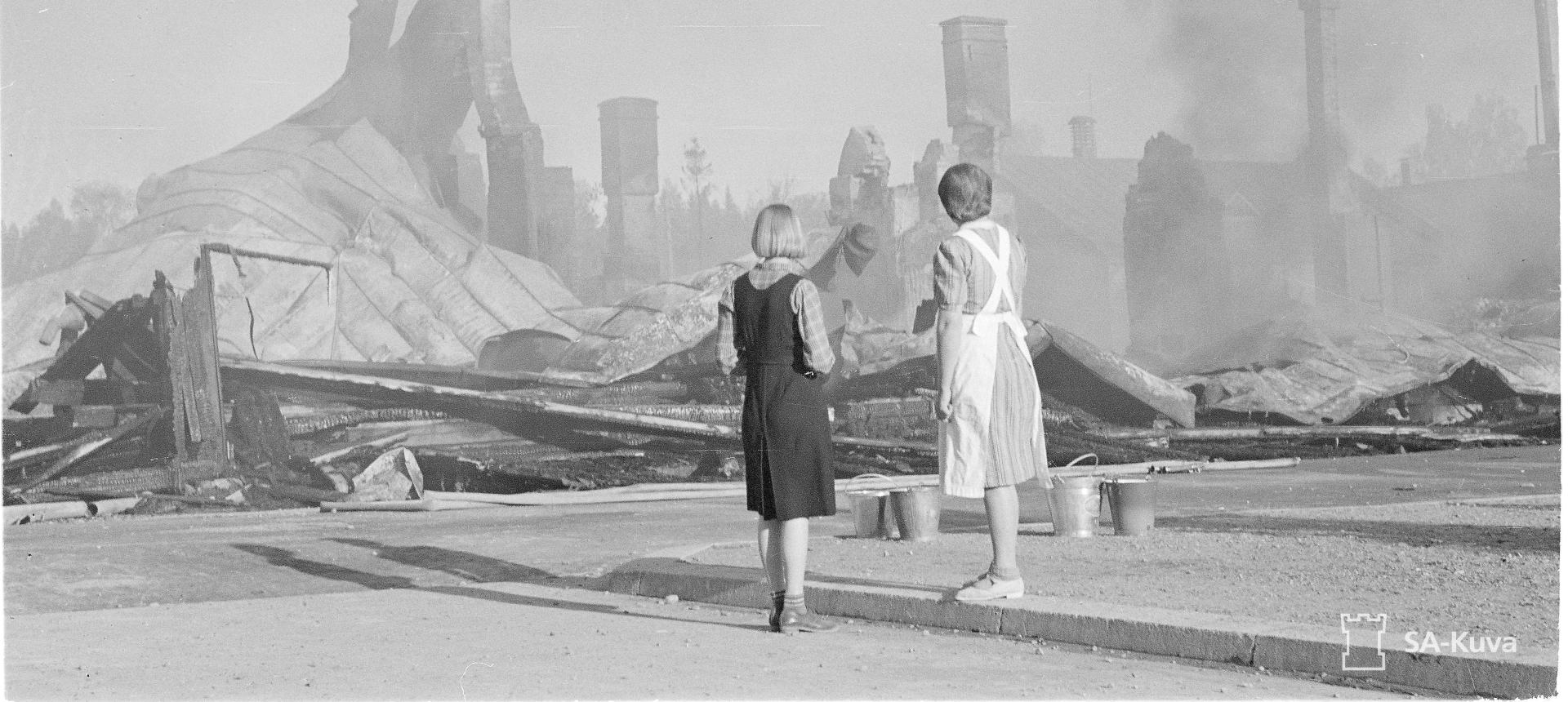 Kaksi naista katselee pommin tuhoamia rakennuksia. 